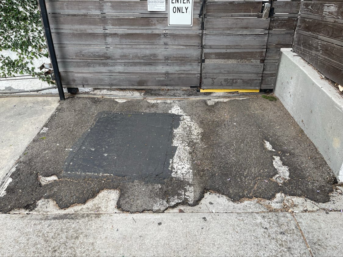 Cracked and patched concrete approach at a gated entry, surface broken and uneven